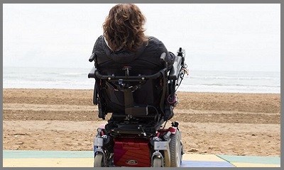 Una donna in sedia a rotelle elettrica, ritratta di spalle, sulla spiaggia, scruta il mare.